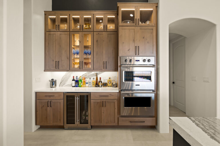 A custom kitchen bar station with warm wood cabinets, glass-front shelving illuminated from within, a built-in wine cooler, and stainless steel double ovens. The countertop displays an array of premium spirits and mixers against a marble backsplash.